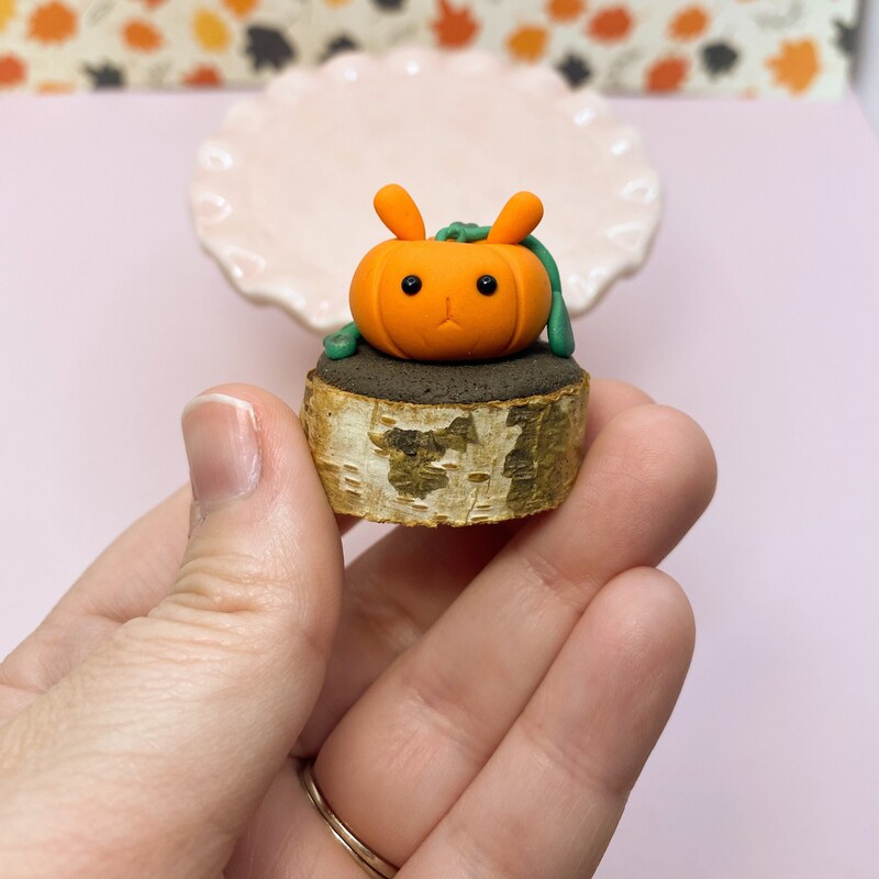 Bunny-shaped pumpkin figurine, orange with perky ears and vines trailing down the side and back on a brown faux-dirt base on top of a small wood slice. Held in a hand with a pale pink background. 