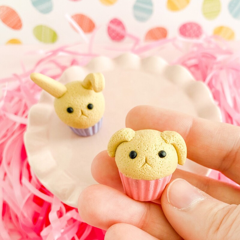 Small bunny cupcake figurine with pink base, vanilla "cake", and lop ears held in a hand. Another bunny cupcake, vanilla with a purple base and perky ears, is in the background sitting on a small, pale pink cake plate with pink easter grass surrounding it. A hint of easter egg patterned background can be seen.