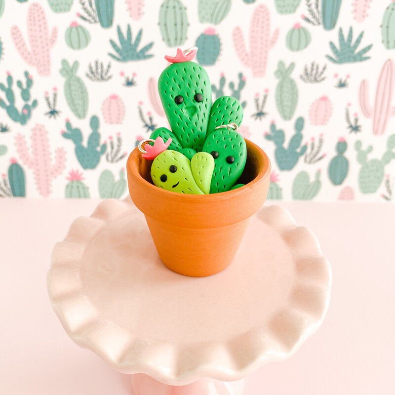 A trio of green cactus charms perched in a terra cotta pot. The tallest is in the back, bright green with a happy :D expression and a pink flower atop their head. The medium cactus is partially hidden but is also bright green with eyes but no mouth. The smallest cactus is in the front, a lighter spring green, with a smile and a flower topper, too. The mini pot sits atop a pale pink cake plate against a pink and green cactus patterned background.