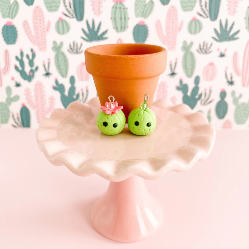 Two spring green barrel shaped cactus charms sit in front of a terra cotta pot on a small, pale pink cake plate. The cactus on the left has a flower atop their head while both have black eyes, no mouth, and a sparkling crystal. The pedestal plate rests on a pink ground against a background of pink and green cacti.