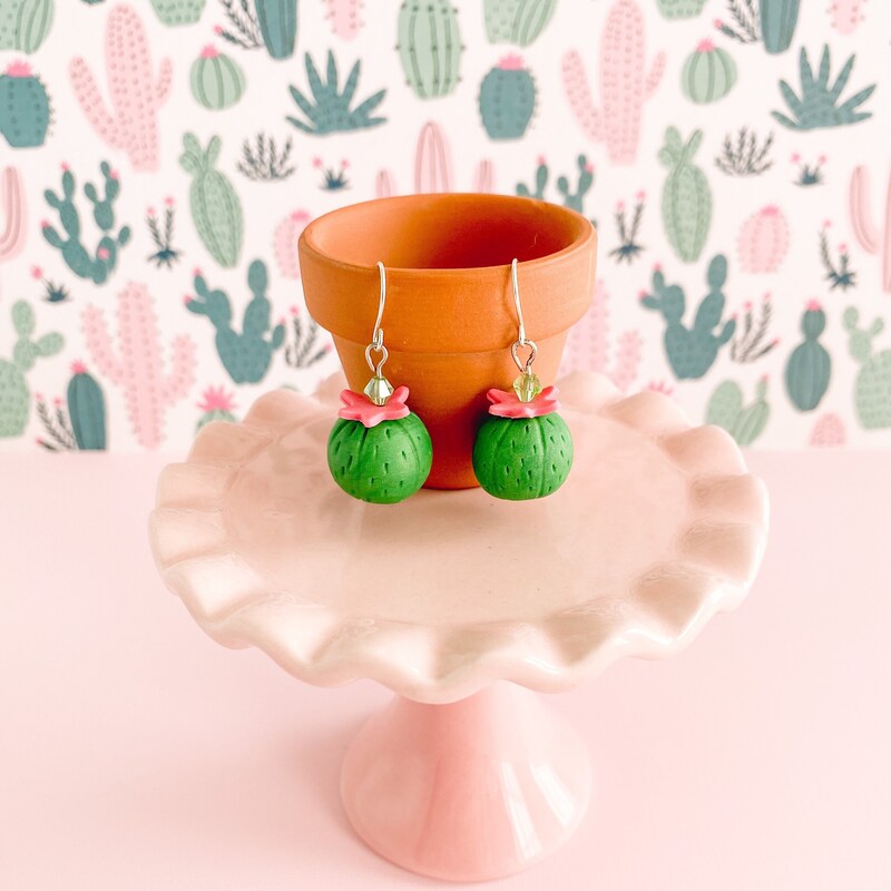 A pair of barrel cactus earrings dangle from silver ear wires hanging on the rim of a small pot. The cacti are bright green spheres with a pink flower and a sparkling green crystal on top. They hang on a mini terra cotta pot sitting on a small pale pink cake plate, on a pink ground and in front of a green and pink cactus patterned background.